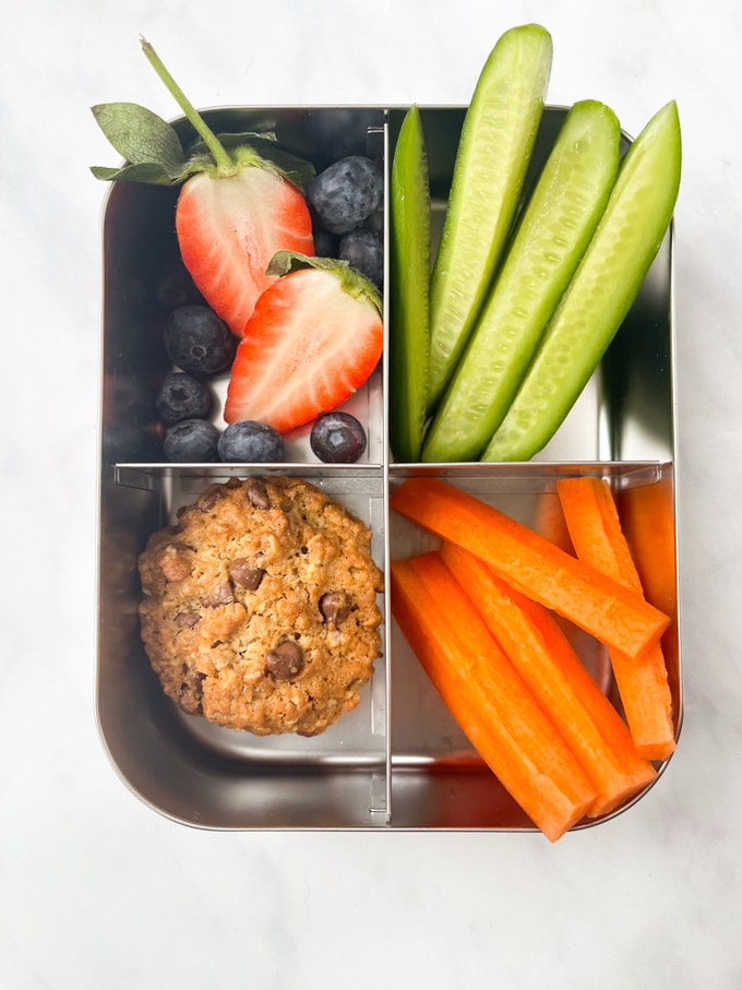 Chunky Chocolate Chip Oat Cookies in a stainless steel compartment lunchbox with cucumber and carrot sticks and blueberries and strawberry