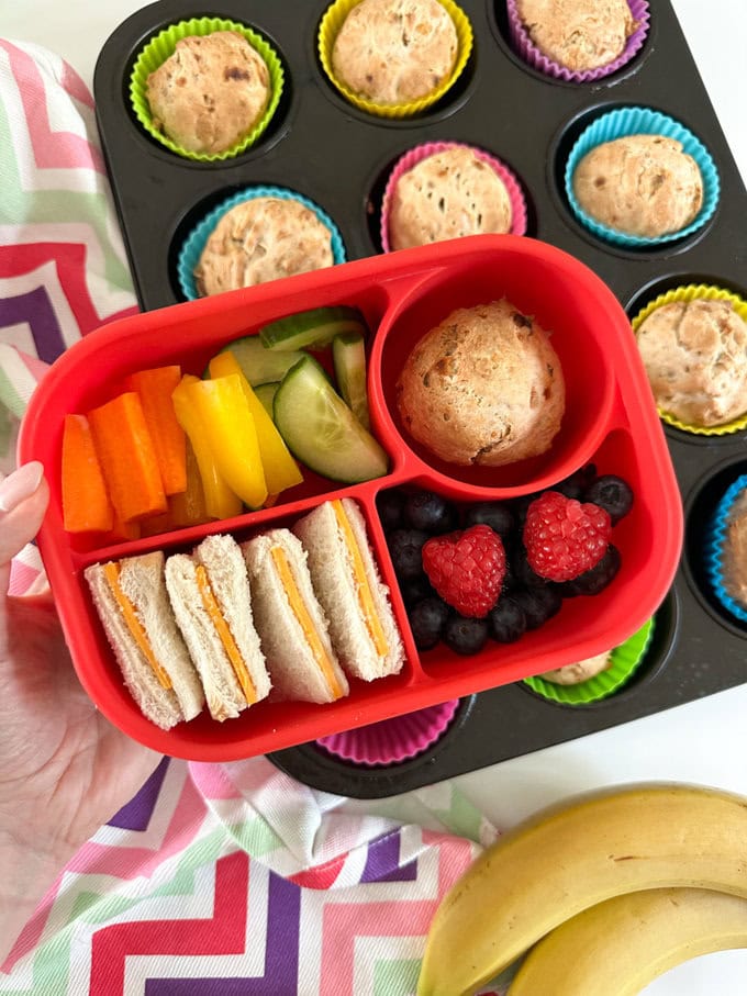 3 Ingredient Banana Muffins presented in a bright red Bento style lunchbox, with cheese sandwiches and fresh berries, cucumber slices and chopped yellow pepper and carrot's.