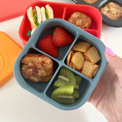 Mini Raspberry & White Chocolate Banana Breads in a blue silicone snack box with mini cheddars, chopped kiwi fruit and a chopped strawberrry.